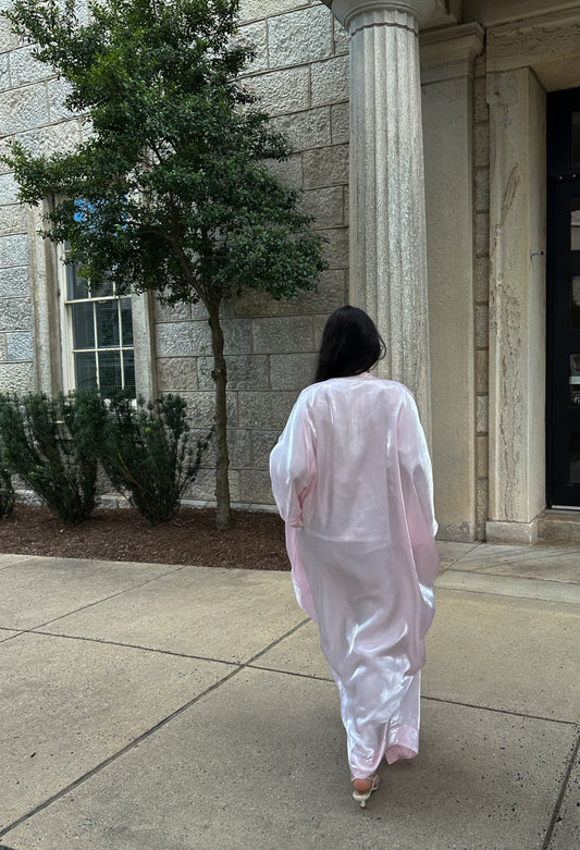 Person in a light pink dress walking on a sidewalk in front of a building with columns.