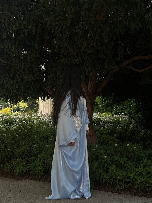 Woman in a blue and white abaya standing outdoors with greenery in the background