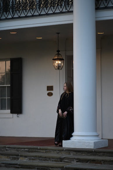 Woman in a black abaya standing on a porch with white columns and a hanging light fixture.