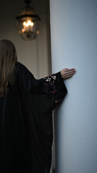 Person in a dark abaya leaning against a white wall with a dimly lit lantern in the background.