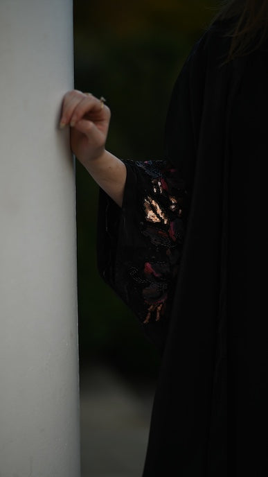 Person wearing a dark abaya with floral patterns leaning against a white wall.