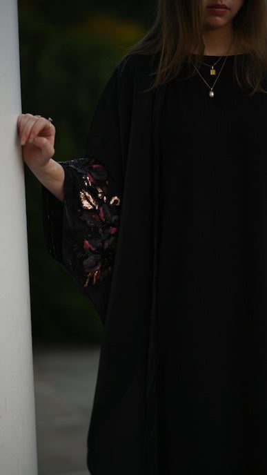 Person wearing a black outfit with floral embroidery, standing against a dark background.