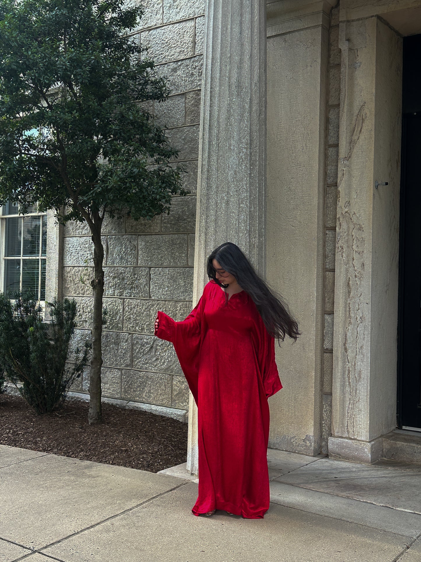 Person wearing a red abaya standing outside a building with stone walls and a tree.