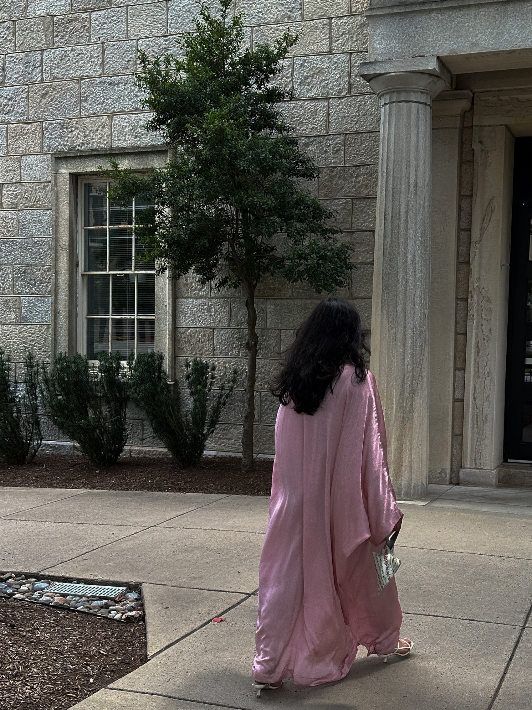 Person in a pink dress walking past a stone building with columns