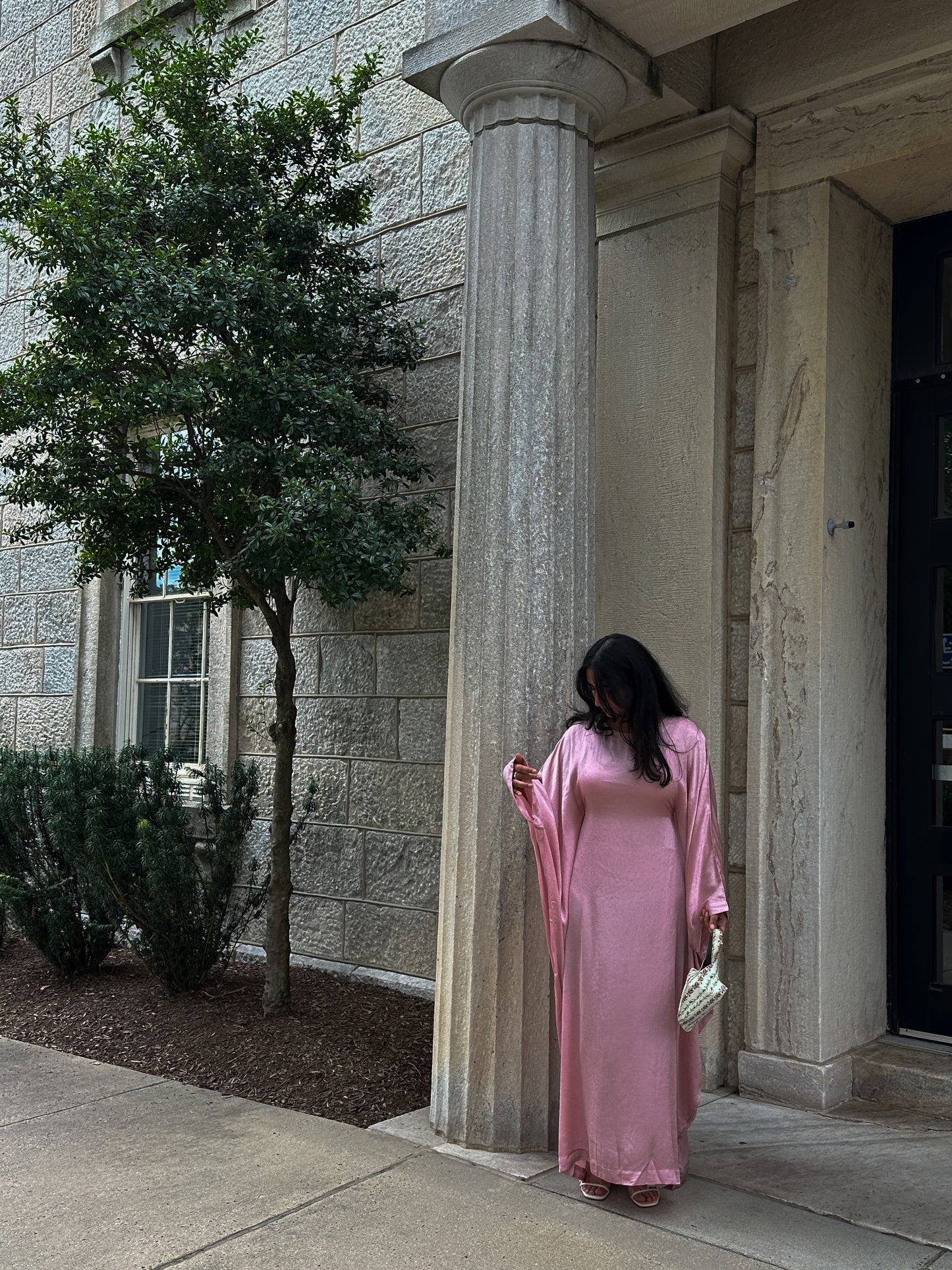 Woman in a pink dress standing next to a stone column outdoors.