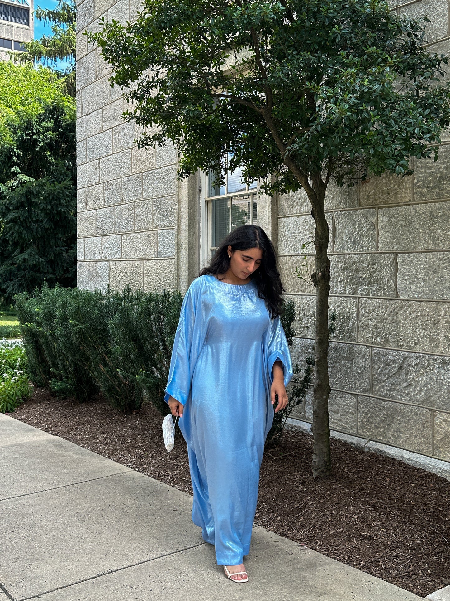 Woman in a blue dress standing on a sidewalk next to a building with trees.
