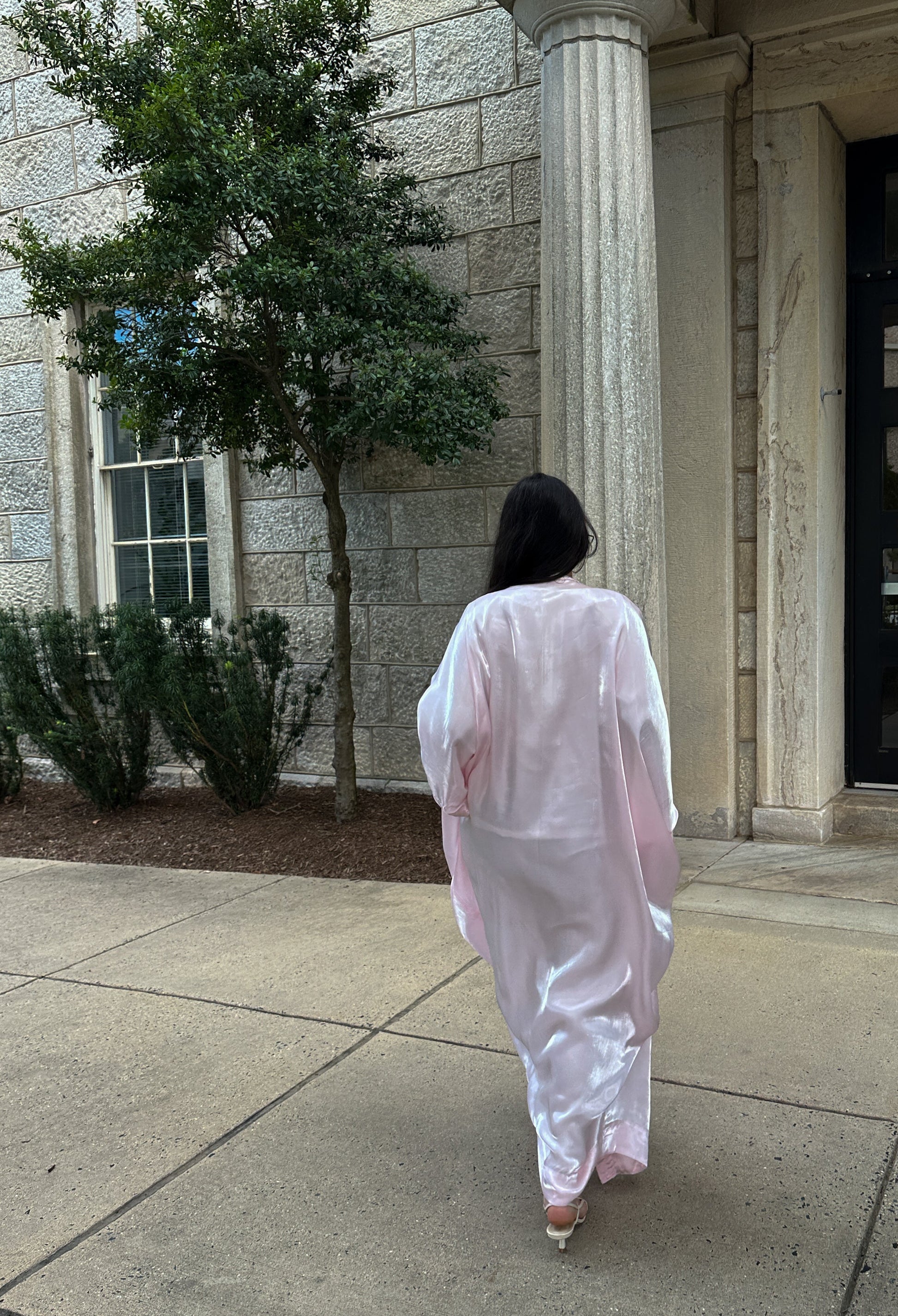Person in a light pink dress walking on a sidewalk in front of a building with columns.