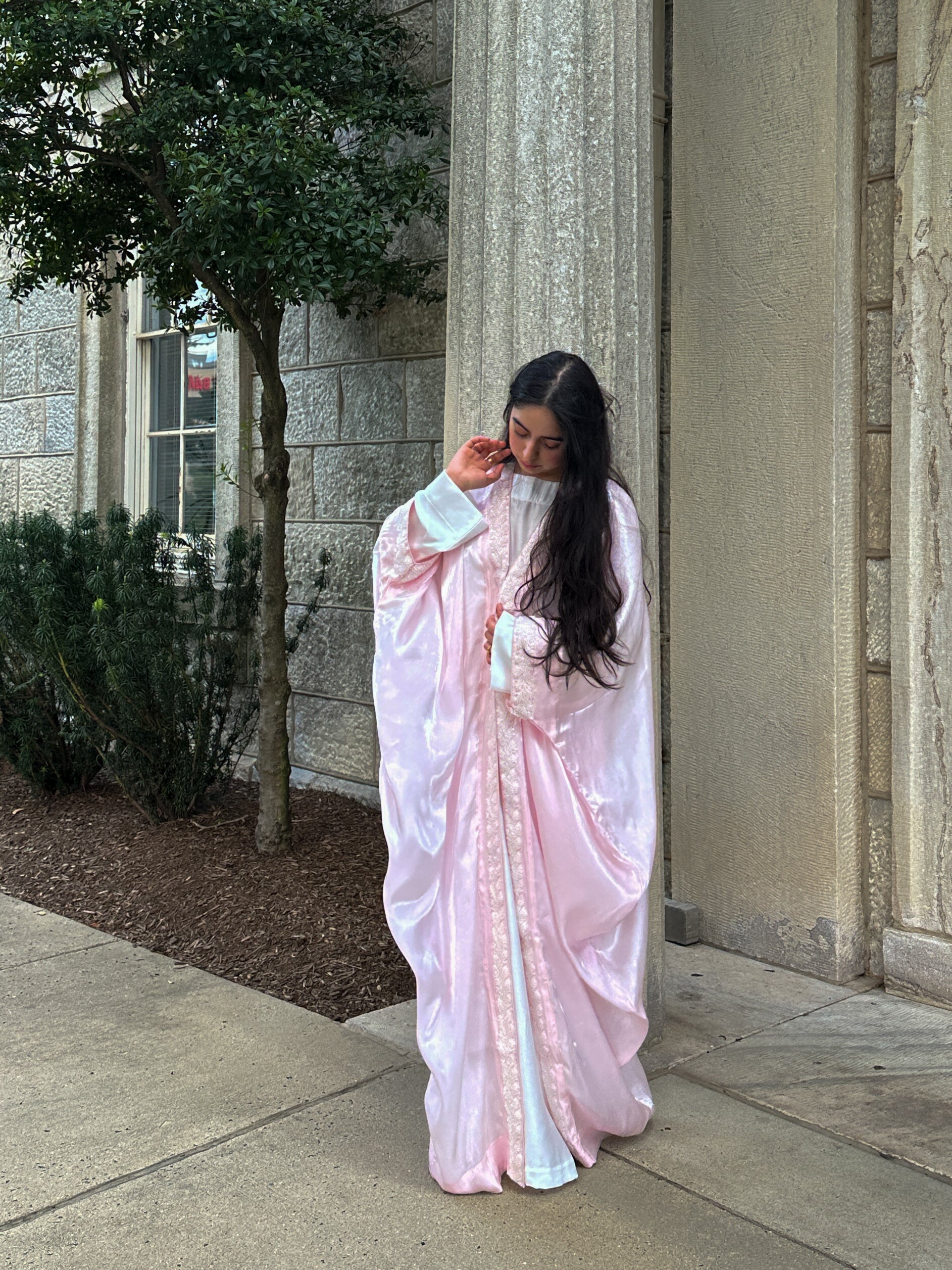 Woman in a light pink abaya standing outdoors near a building.
