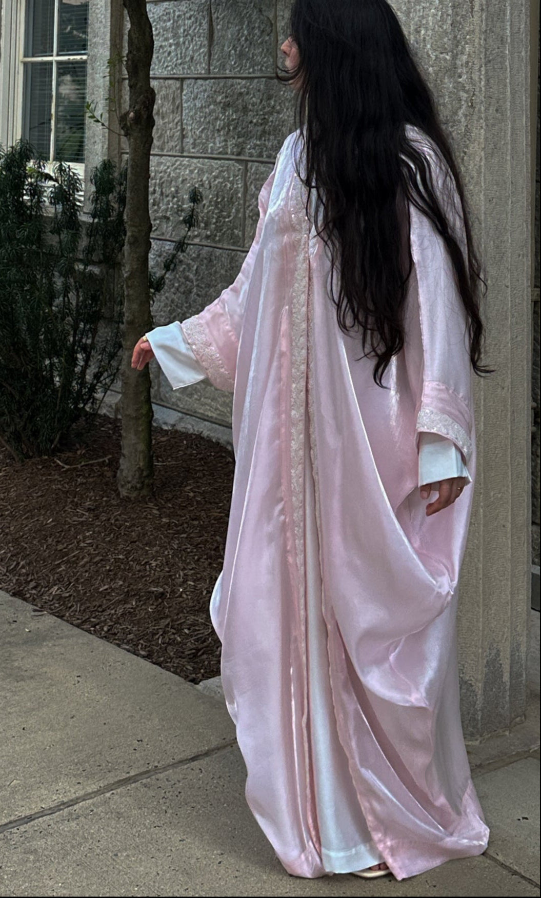Woman in a light pink dress standing next to a stone building with trees and bushes in the background.