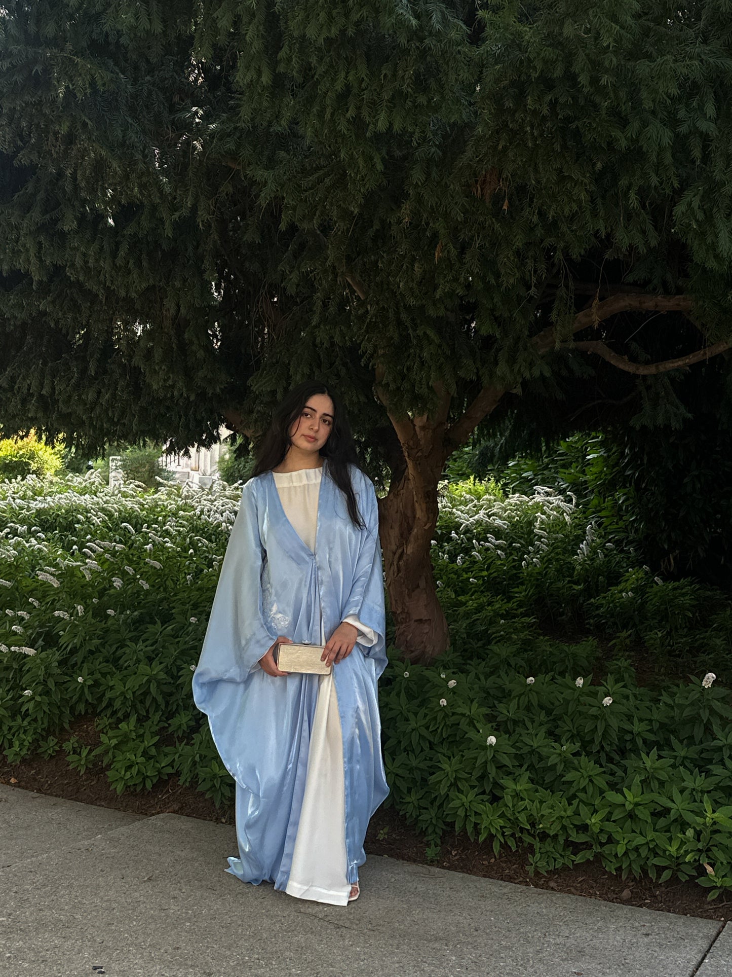 Woman in a blue and white abaya standing outdoors with greenery in the background