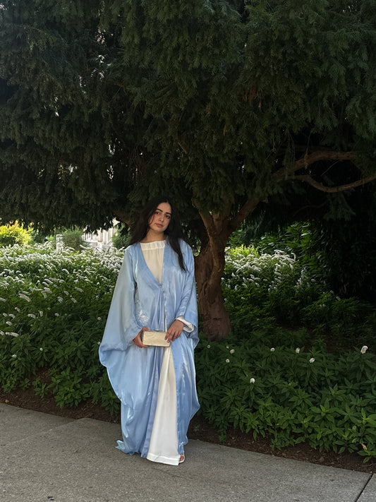 Woman in a blue and white abaya standing outdoors with greenery in the background
