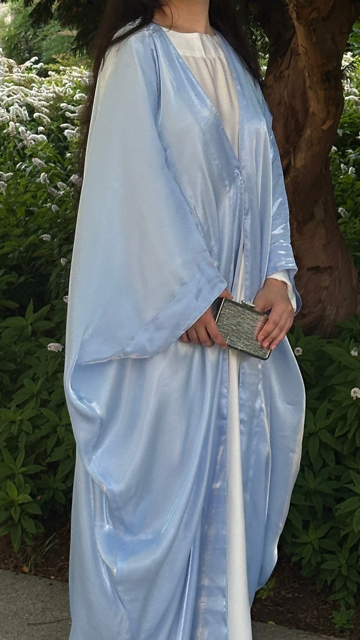 Woman in a blue and white abaya standing outdoors with greenery in the background