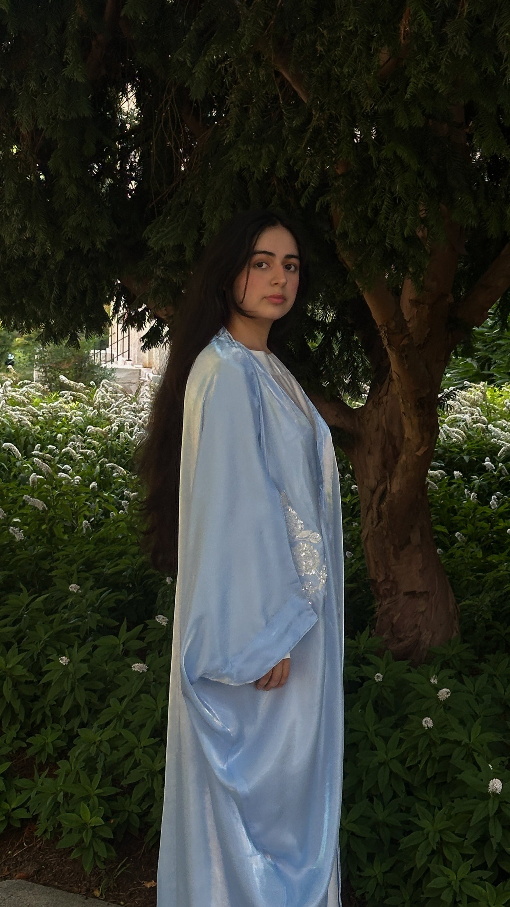 Woman in a blue and white abaya standing outdoors with greenery in the background