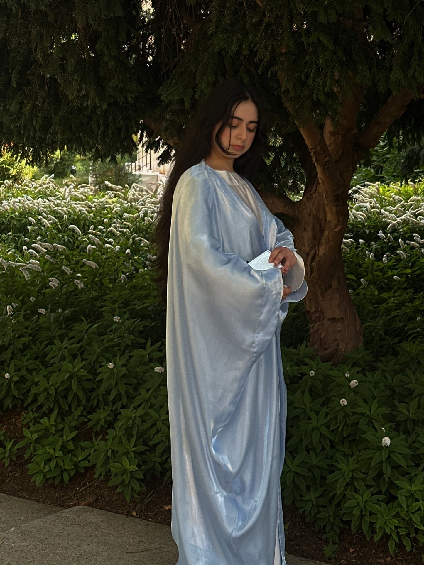 Woman in a blue and white abaya standing outdoors with greenery in the background