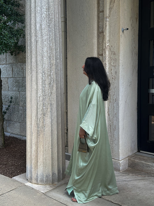 Woman in a long, sage abaya standing in front of stone columns.