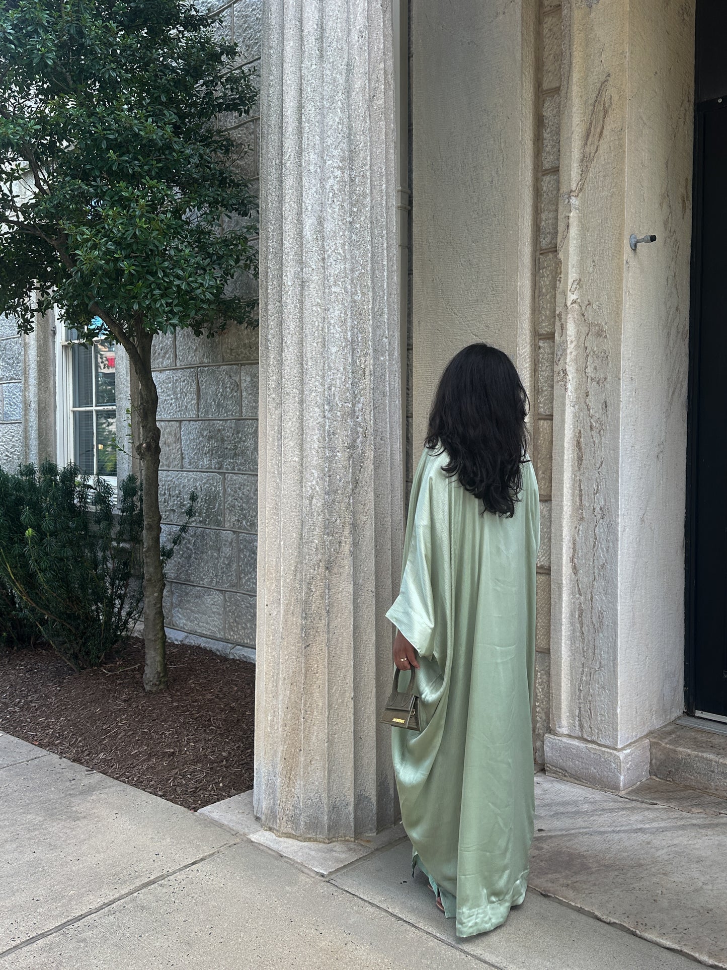 Woman in a long, sage abaya standing in front of stone columns.
