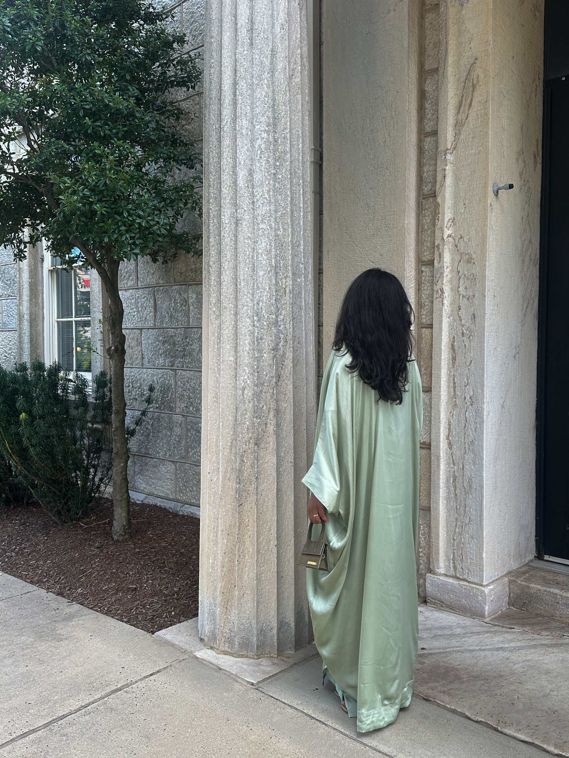 Woman in a long, sage abaya standing in front of stone columns.