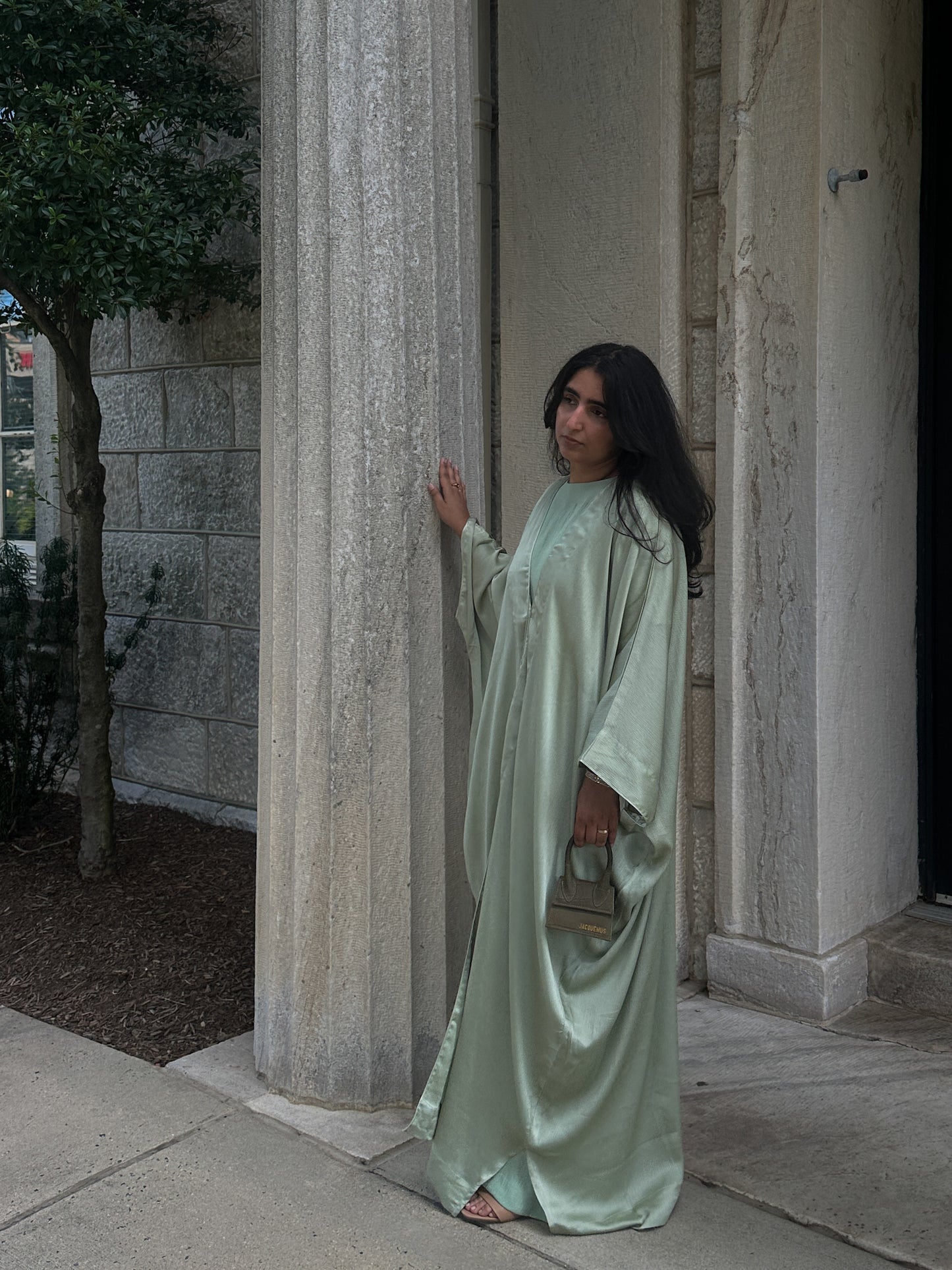 Woman in a long, sage abaya standing in front of stone columns.