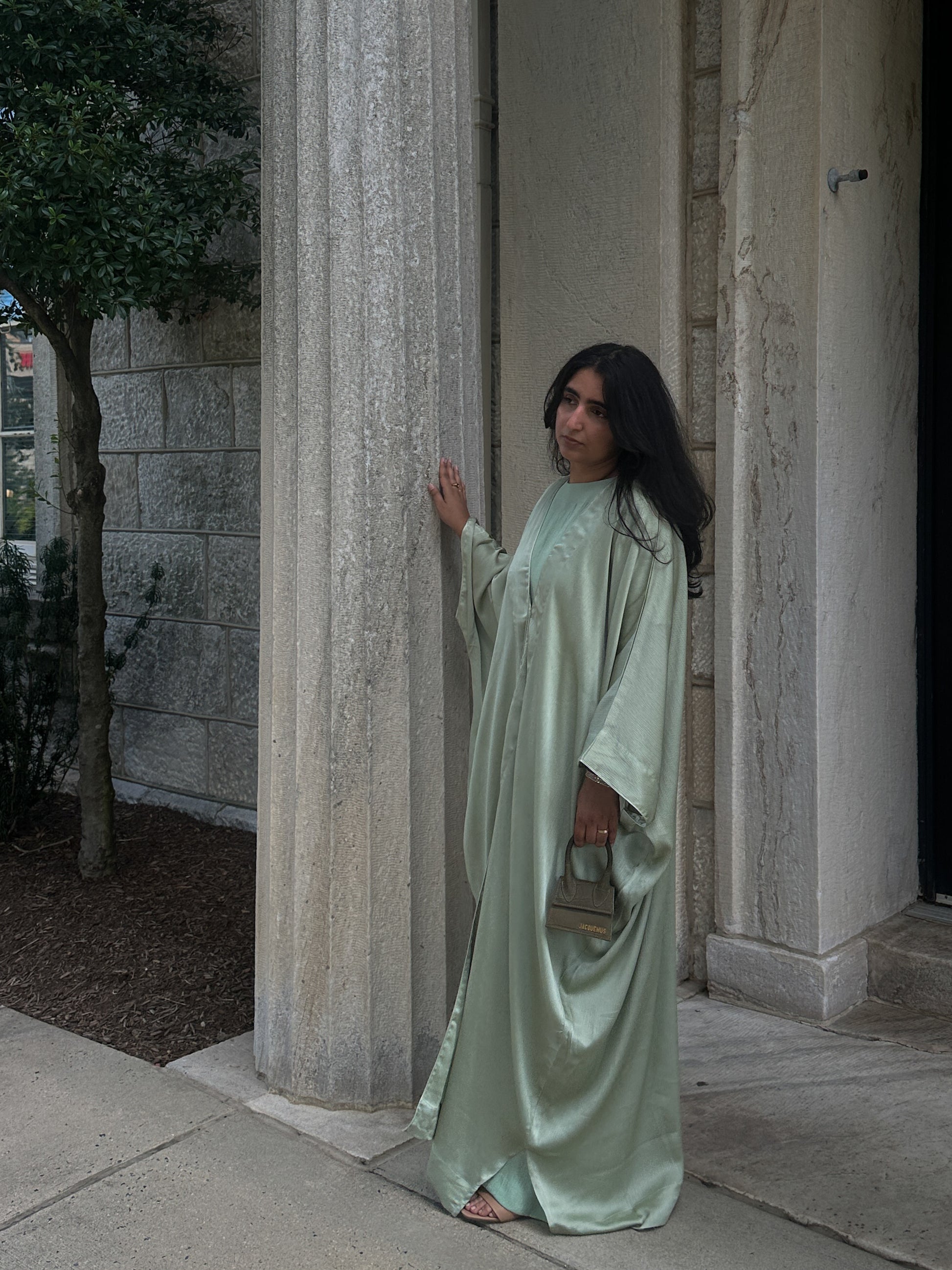 Woman in a long, sage abaya standing in front of stone columns.
