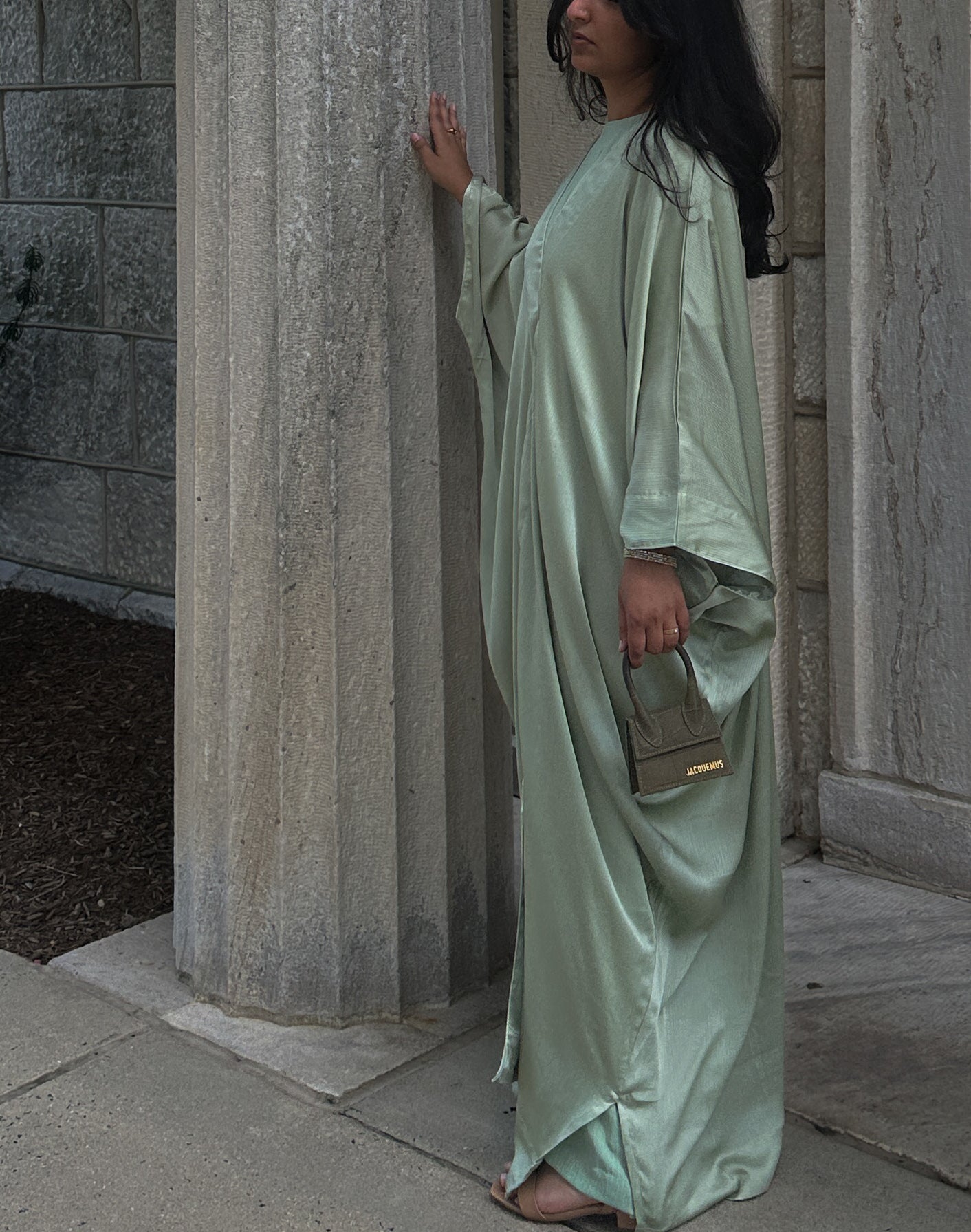 Woman in a long, sage abaya standing in front of stone columns.