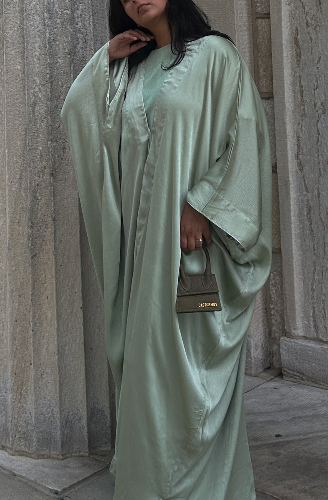 Woman in a Sage Abaya standing against a stone building with columns.