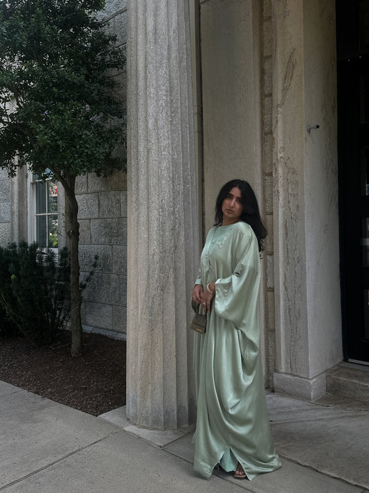 Woman in a long, sage abaya standing in front of stone columns.