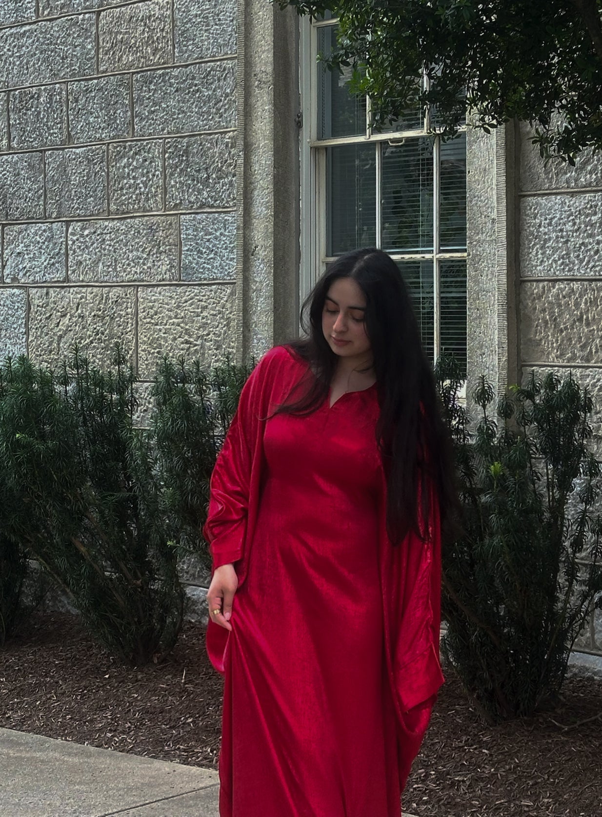 Woman in a red abaya standing outdoors against a stone wall.